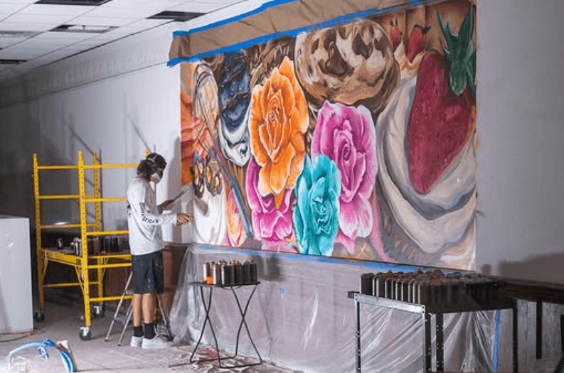 Mural artist Mark Francis painting the Brittany's Cupcake Shoppe bakery mural featuring cupcakes, flowers, and desserts