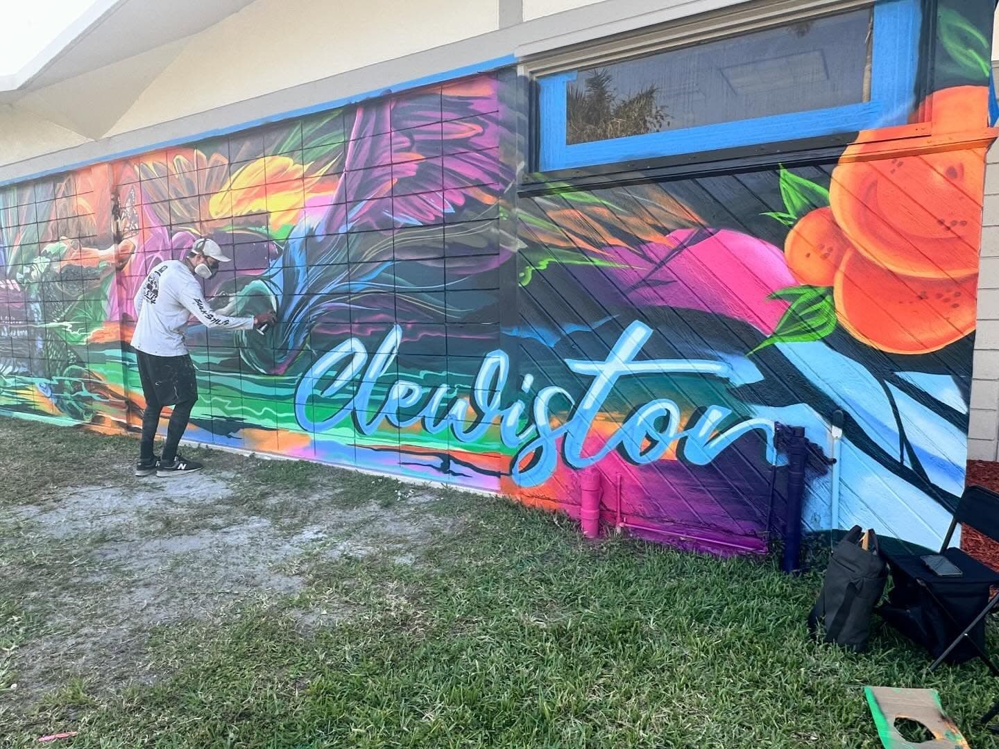 Mark Francis of Bulk Styles spray painting the Clewiston Sugar Festival mural—tropical foliage, Clewiston lettering, and citrus