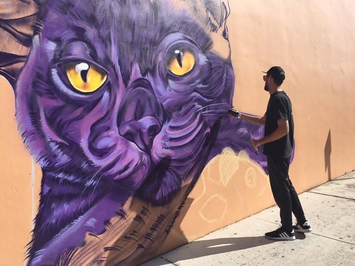 Artist spray painting a stylized purple cat face mural with yellow eyes on an outdoor wall